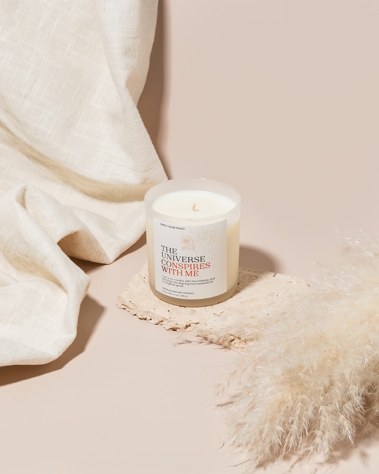 An Ignite Your Peace candle glowing on travertine with linen and dried pampas