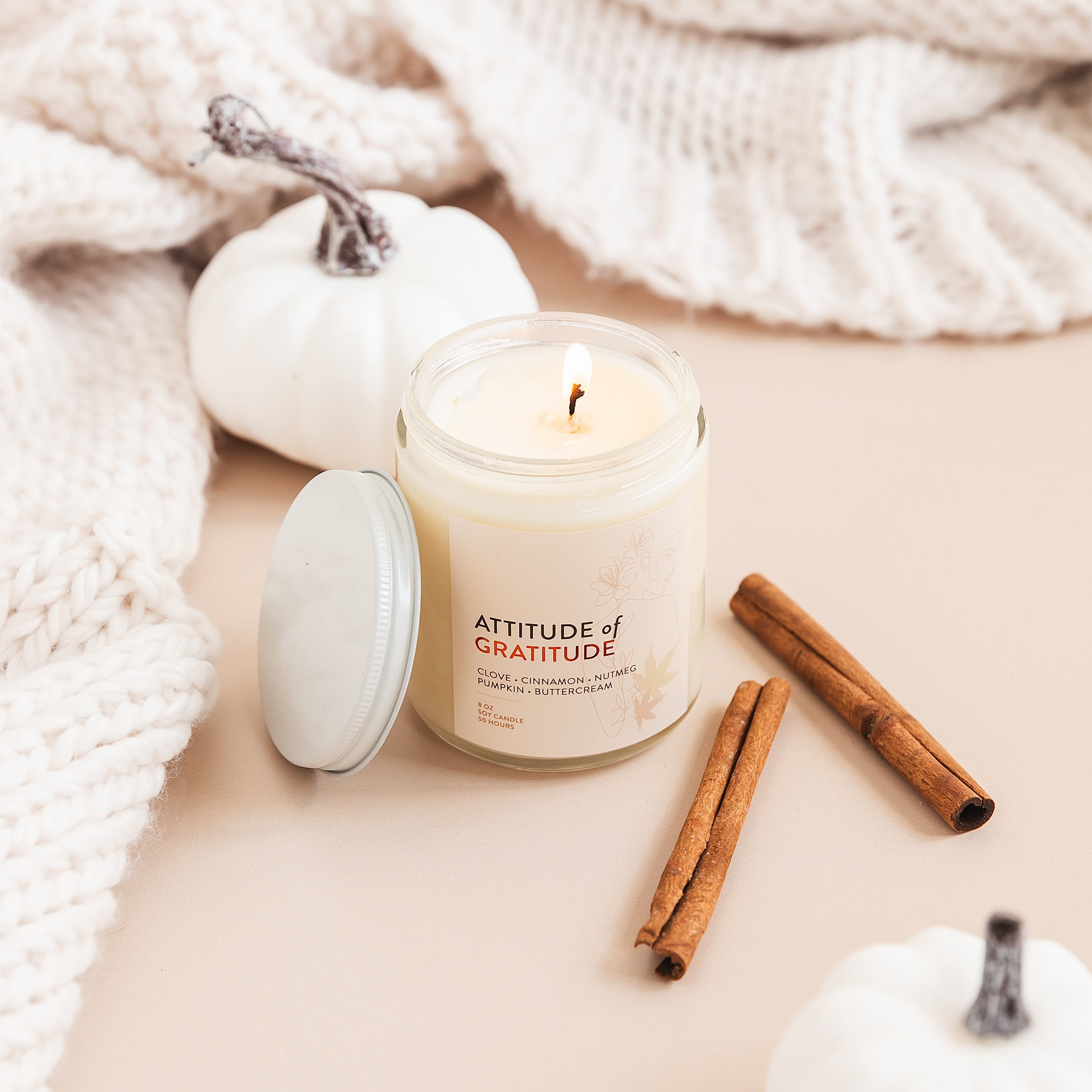 Candle labeled 'Attitude of Gratitude' with cinnamon sticks and pumpkins on a soft surface