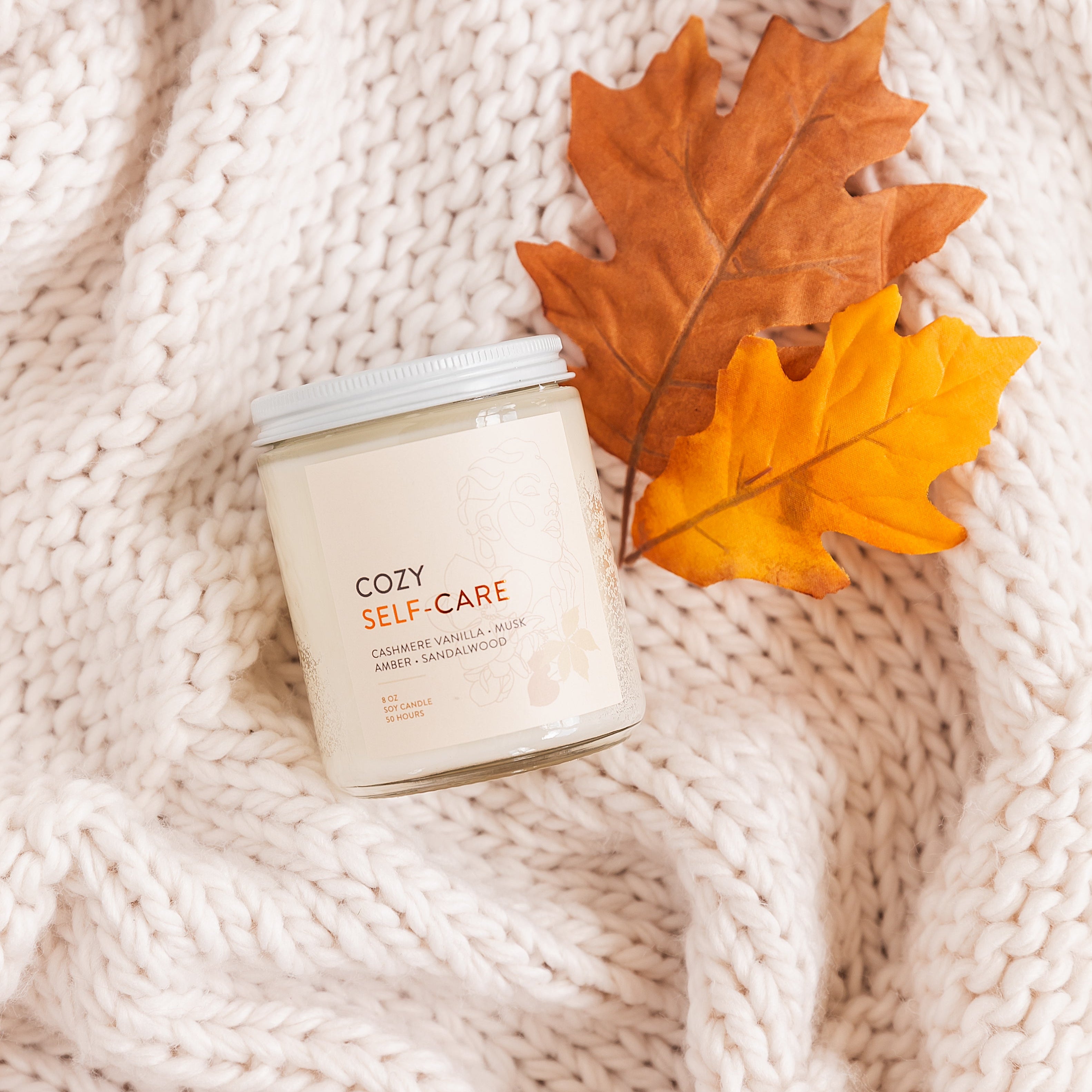 Candle labeled 'Cozy Self-Care' with autumn leaves on a textured surface