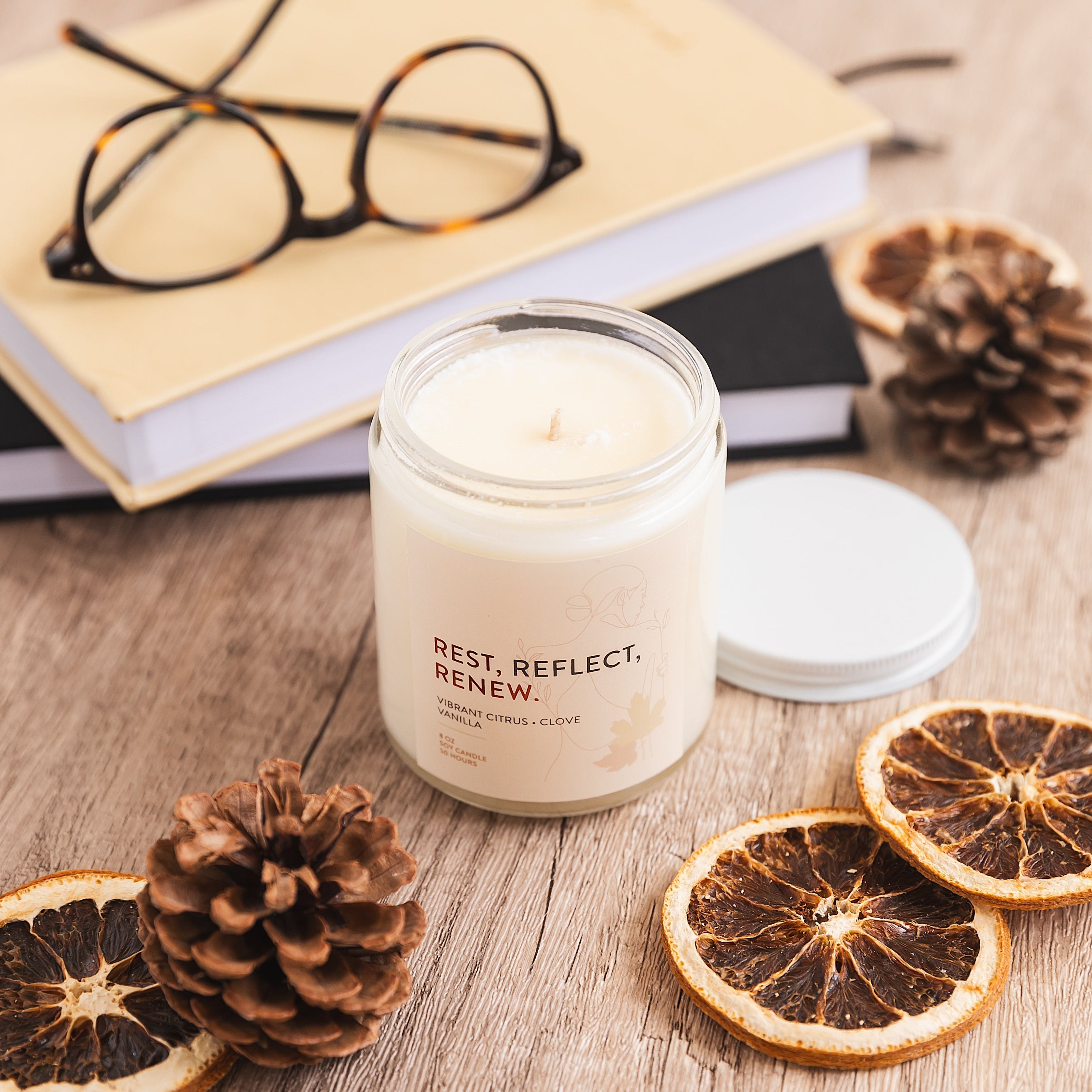 Candle with 'Rest Reflect Renew' text on a wooden surface with dried oranges and pinecones.